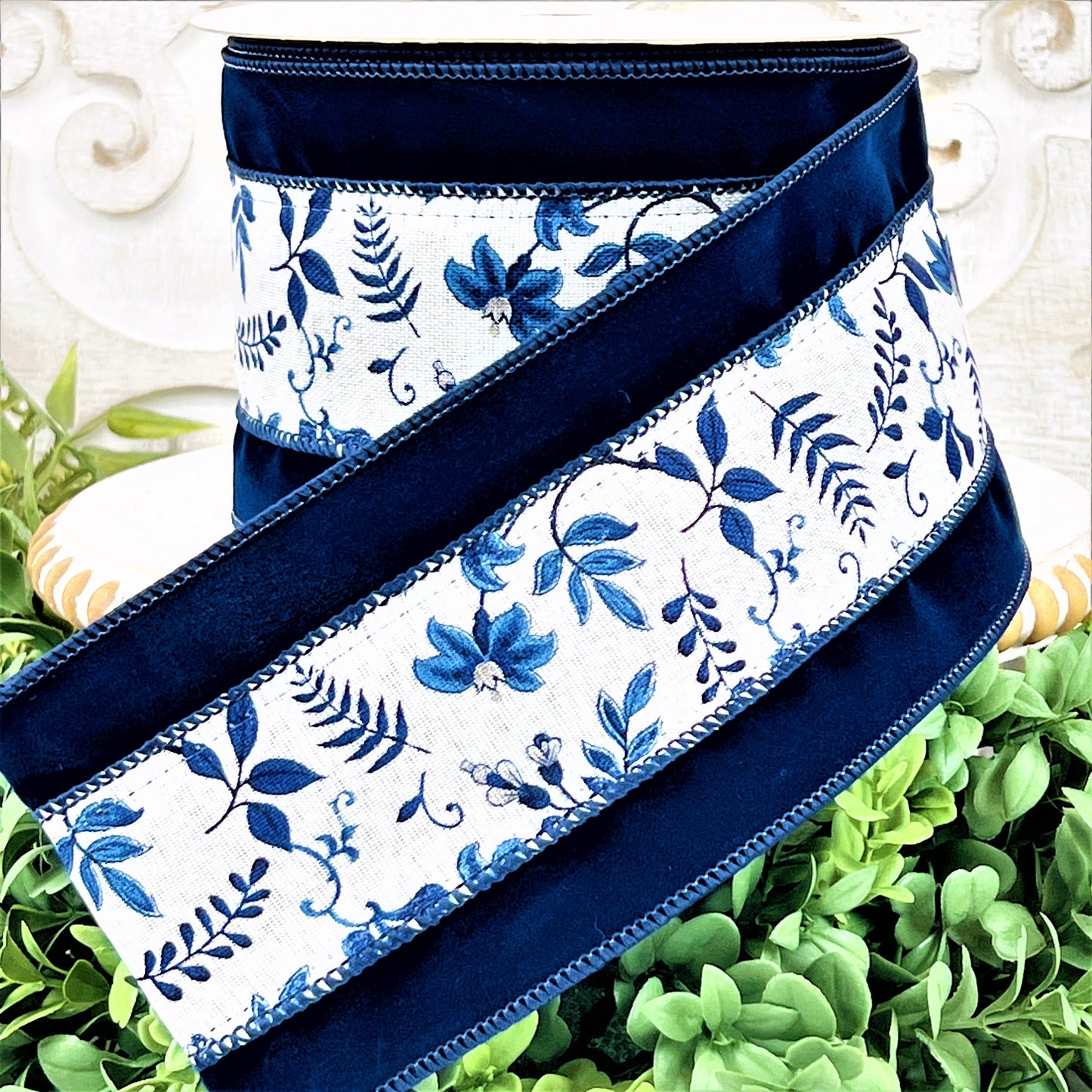 A roll of navy velvet ribbon with a blue and white floral vine in the center sits upon a white wood pedestal surrounded by greenery.