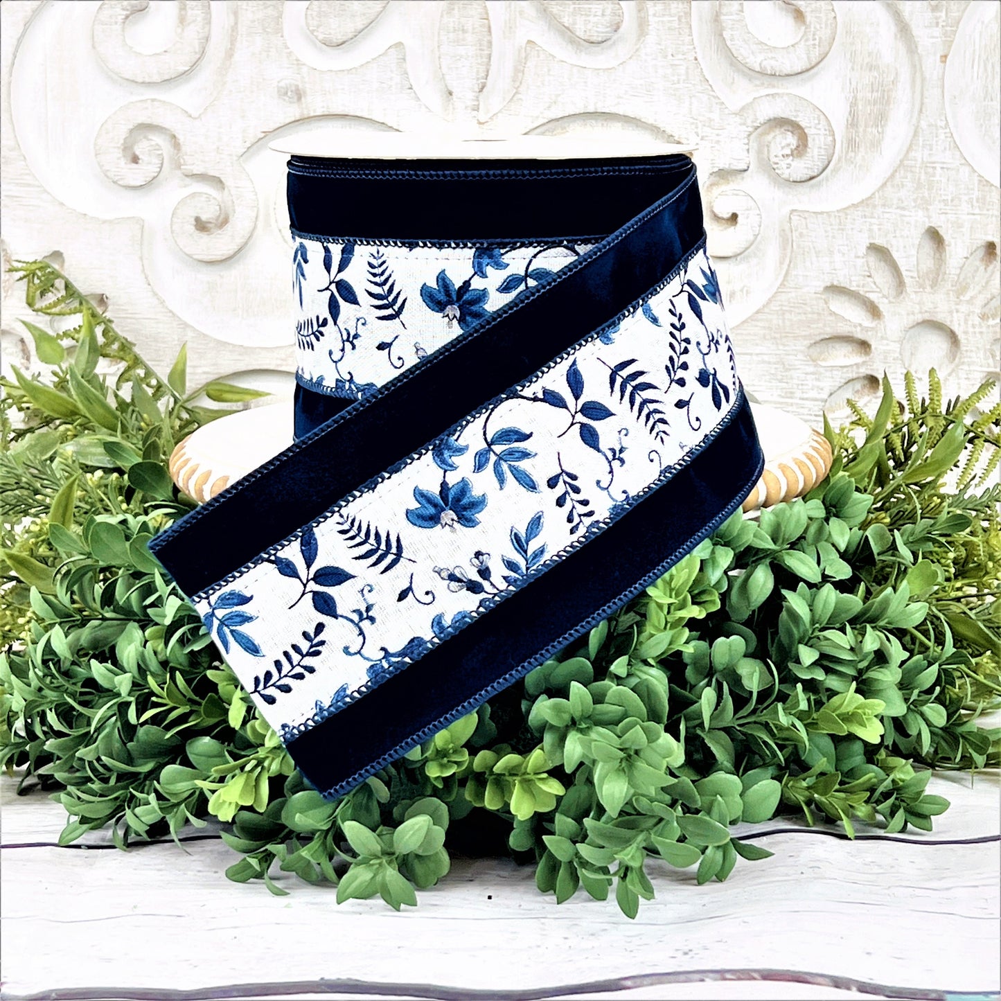 A roll of navy velvet ribbon with a blue and white floral vine in the center sits upon a white wood pedestal surrounded by greenery.