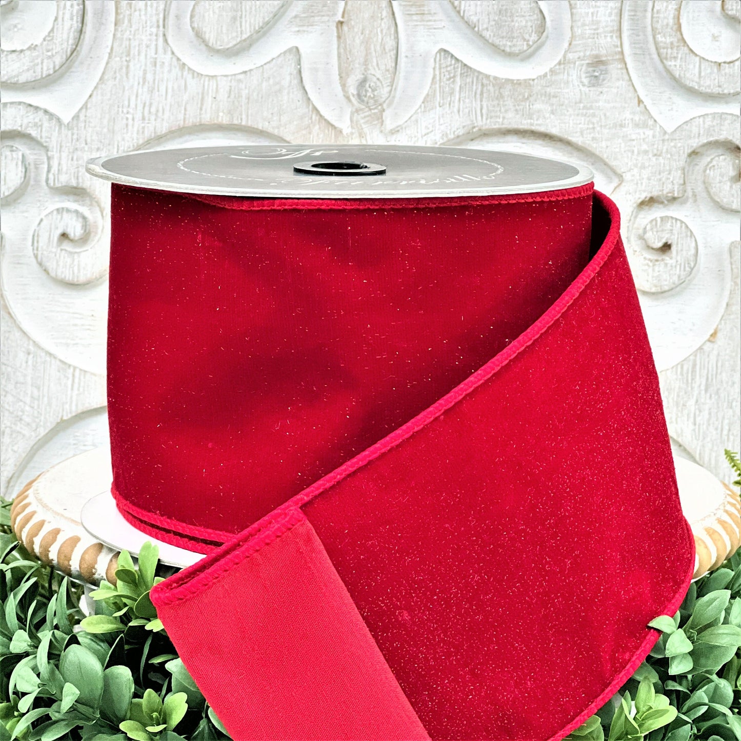 A 4 inch roll of red diamond dust velvet ribbon sits on top of a white wood pedestal surrounded by greenery.