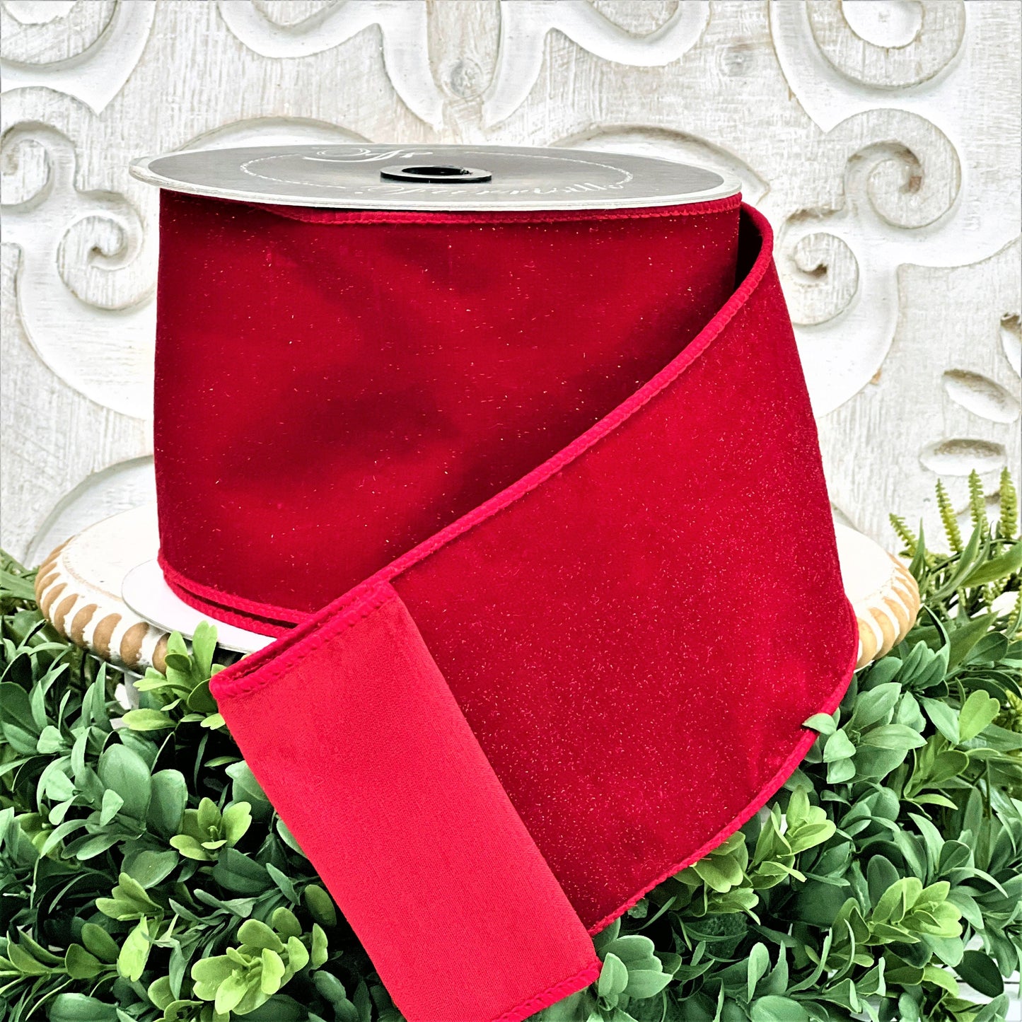 A 4 inch roll of red diamond dust velvet ribbon sits on top of a white wood pedestal surrounded by greenery.
