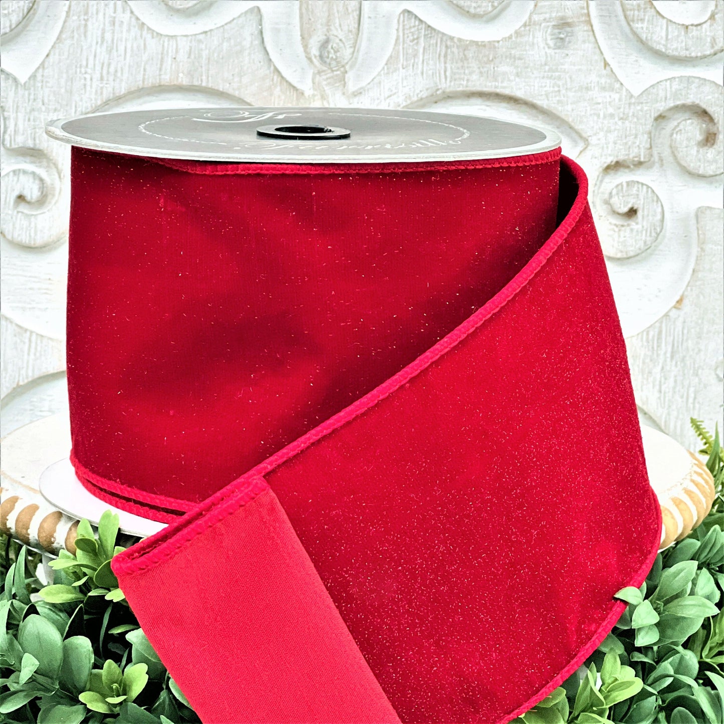 A 4 inch roll of red diamond dust velvet ribbon sits on top of a white wood pedestal surrounded by greenery.