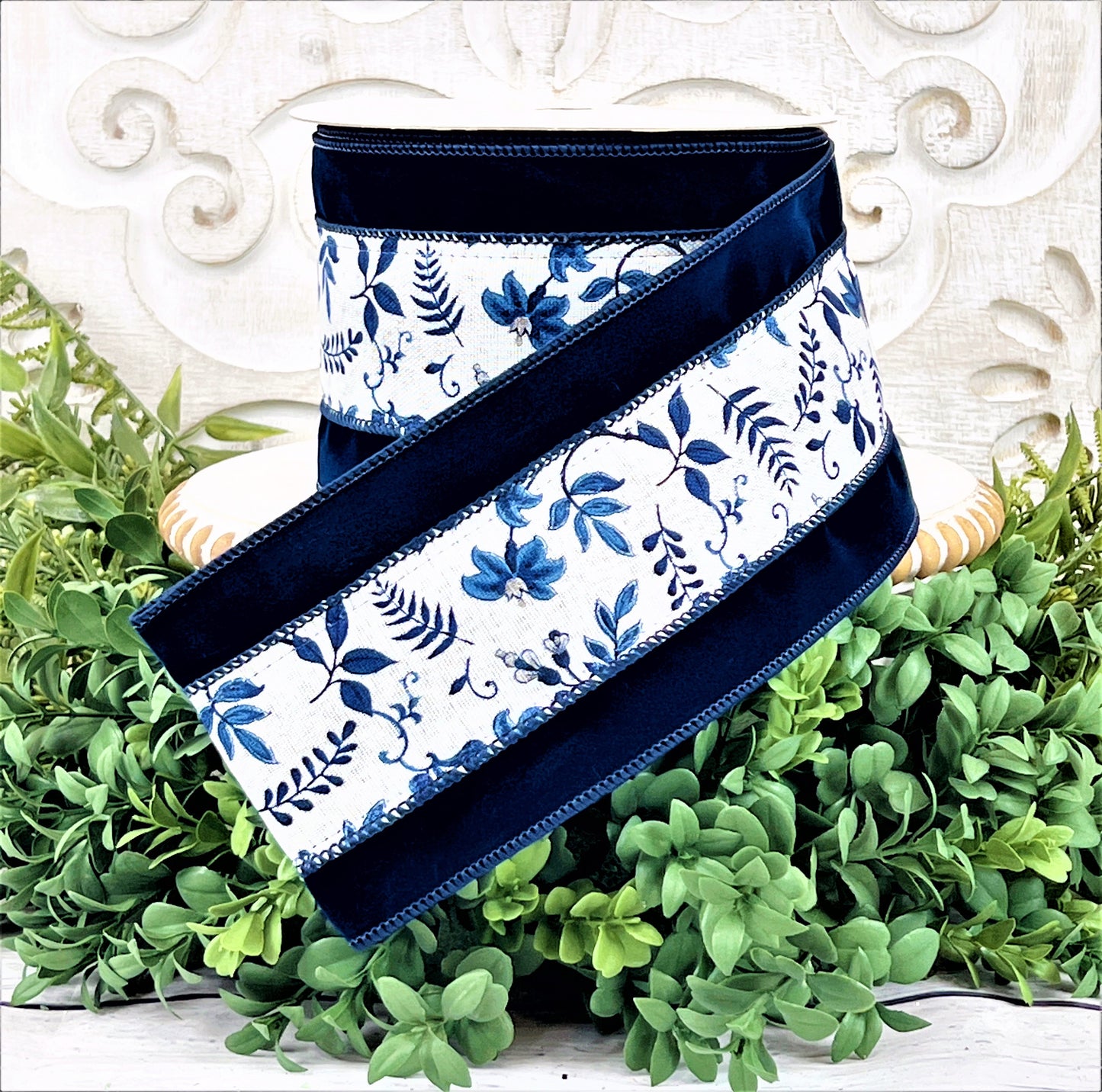 A roll of navy velvet ribbon with a blue and white floral vine in the center sits upon a white wood pedestal surrounded by greenery.