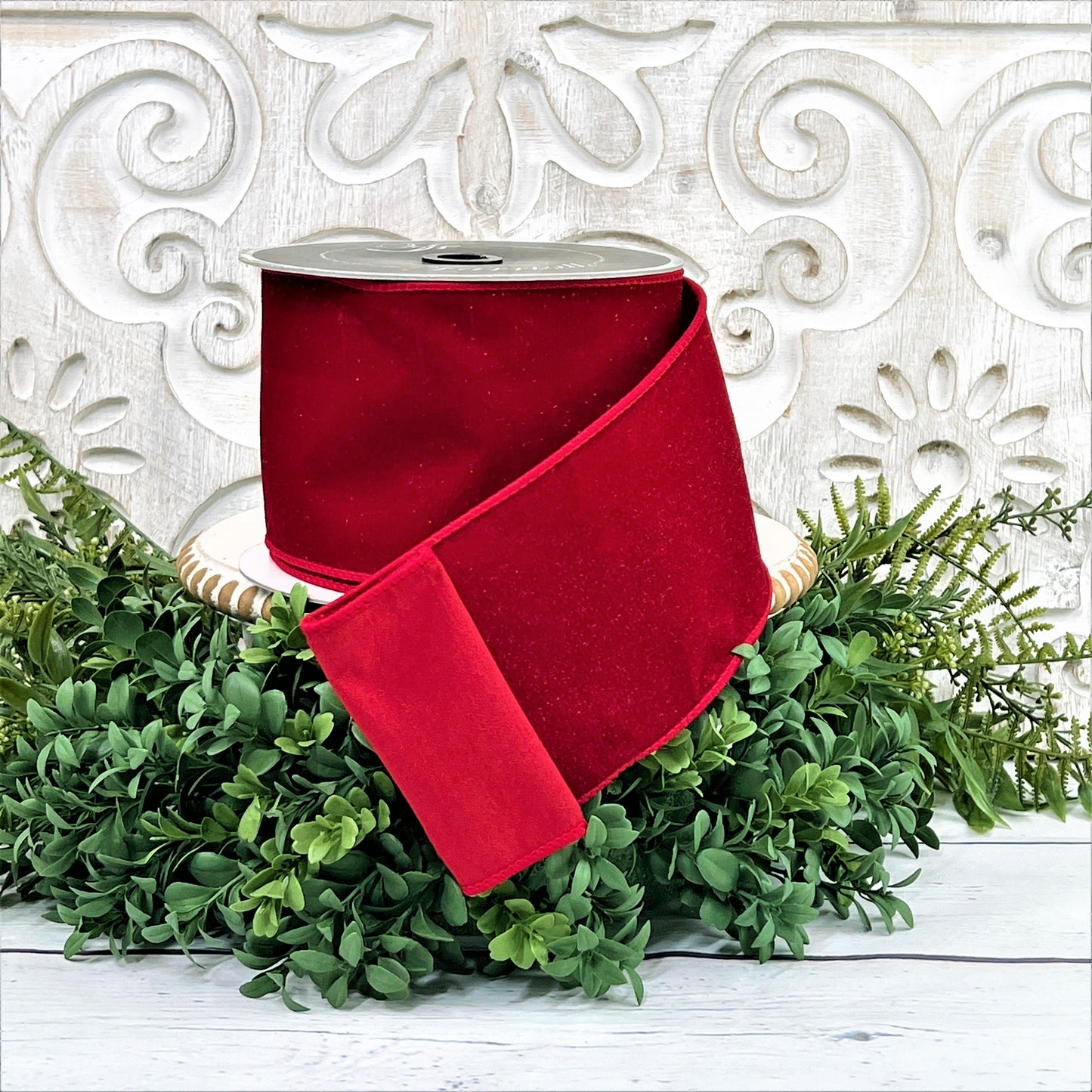 A 4 inch roll of red diamond dust velvet ribbon sits on top of a white wood pedestal surrounded by greenery.