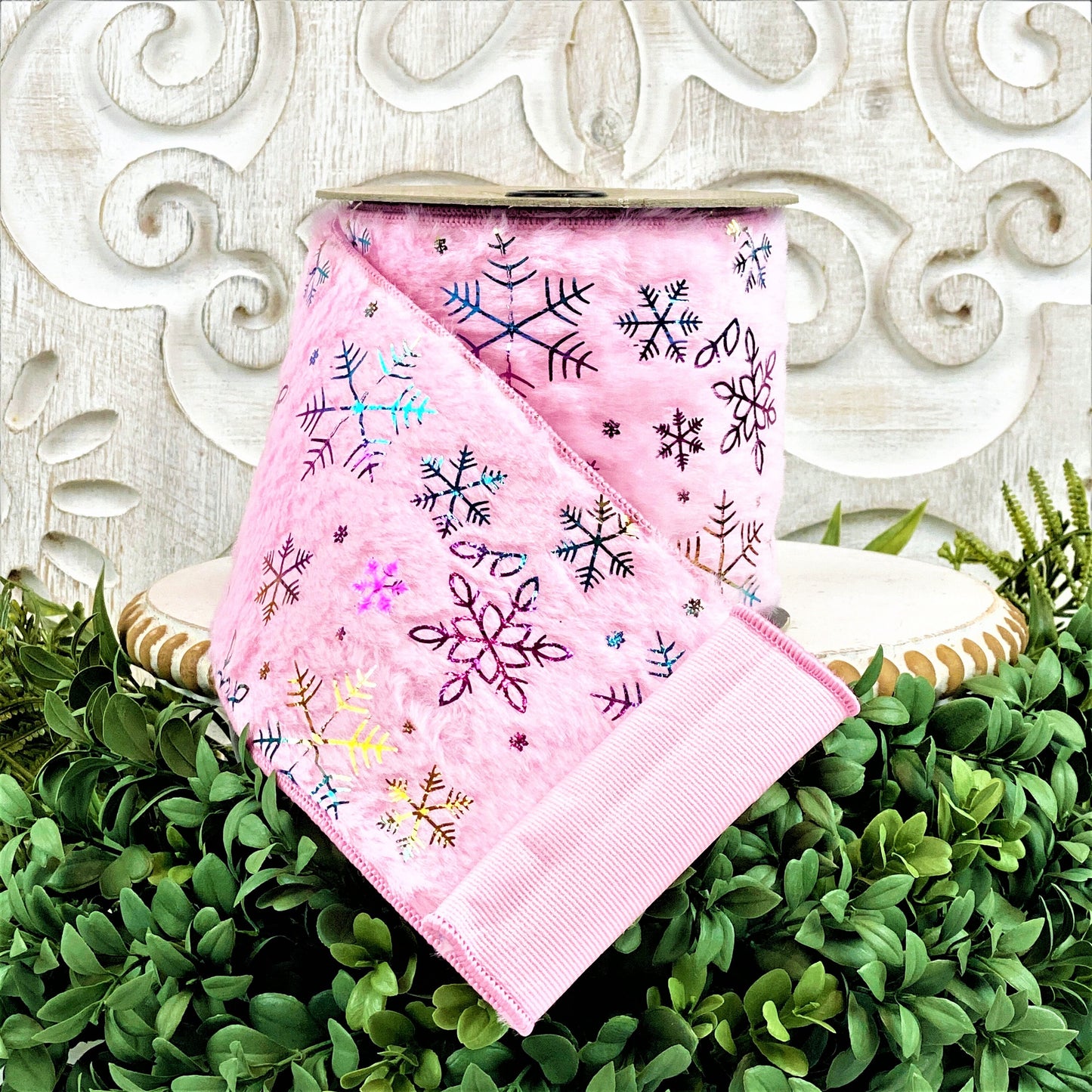 A roll of pink faux fur ribbon sits on a white wood pedestal surrounded by boxwood. The ribbon has irridescent snowflakes all over it, which reflect different colors.