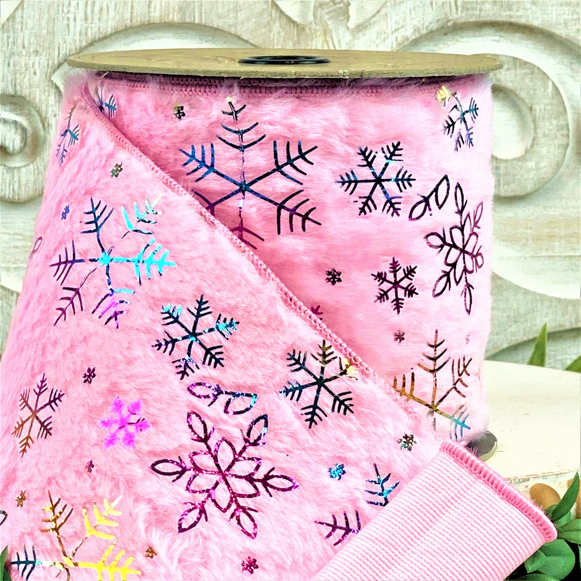 A roll of pink faux fur ribbon sits on a white wood pedestal surrounded by boxwood. The ribbon has irridescent snowflakes all over it, which reflect different colors.