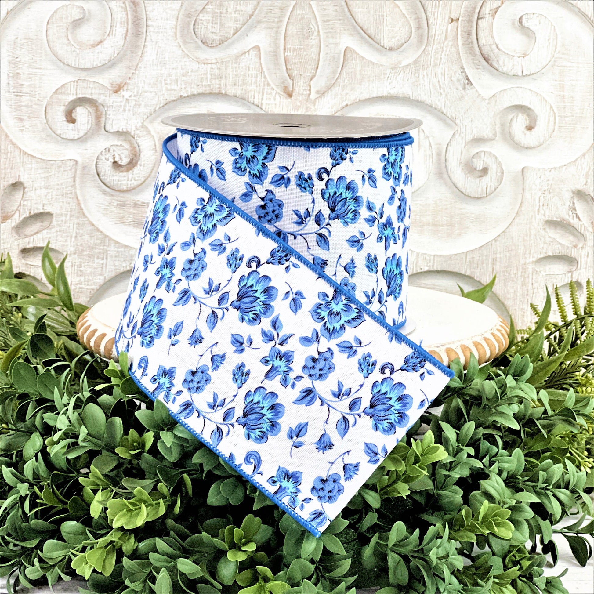 A 4&quot; roll of blue and white Dutch Porcelan flowered ribbon sits on top of a white wood pedestal surrounded by greenery.