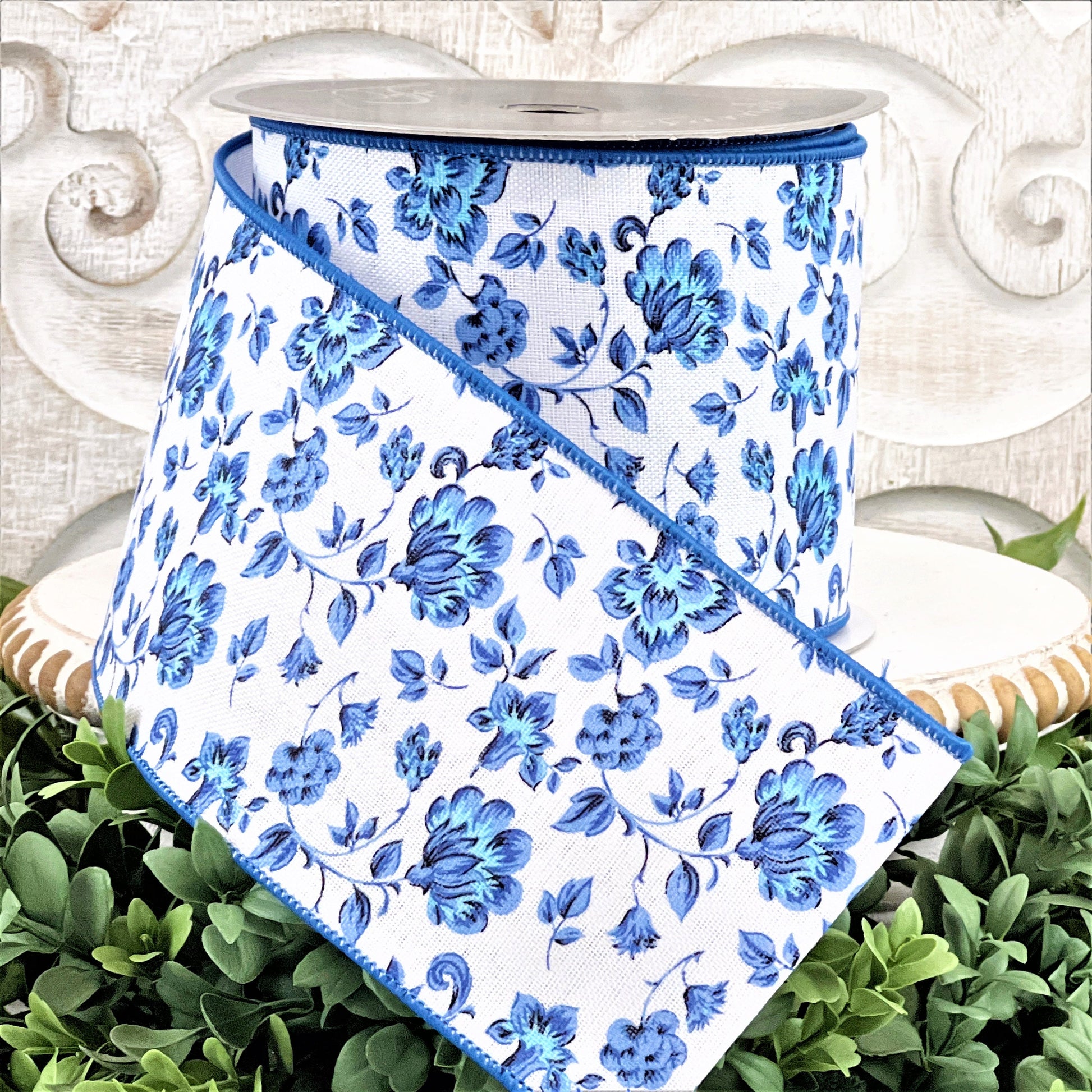 A 4&quot; roll of blue and white Dutch Porcelan flowered ribbon sits on top of a white wood pedestal surrounded by greenery.