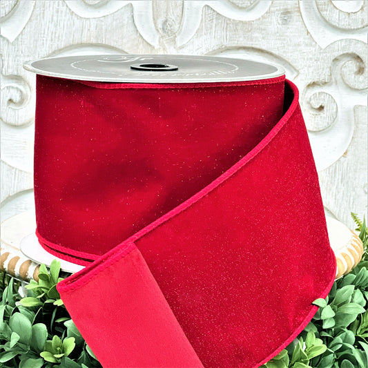 A 4 inch roll of red diamond dust velvet ribbon sits on top of a white wood pedestal surrounded by greenery.