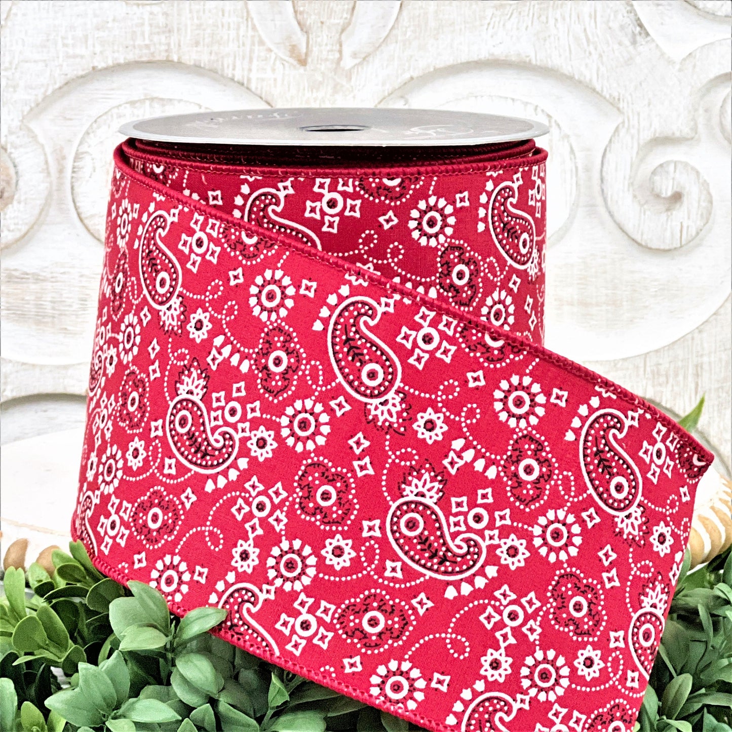 A 4&quot; roll of red paisley print ribbon sits on a white wood pedestal surrounded by greenery.