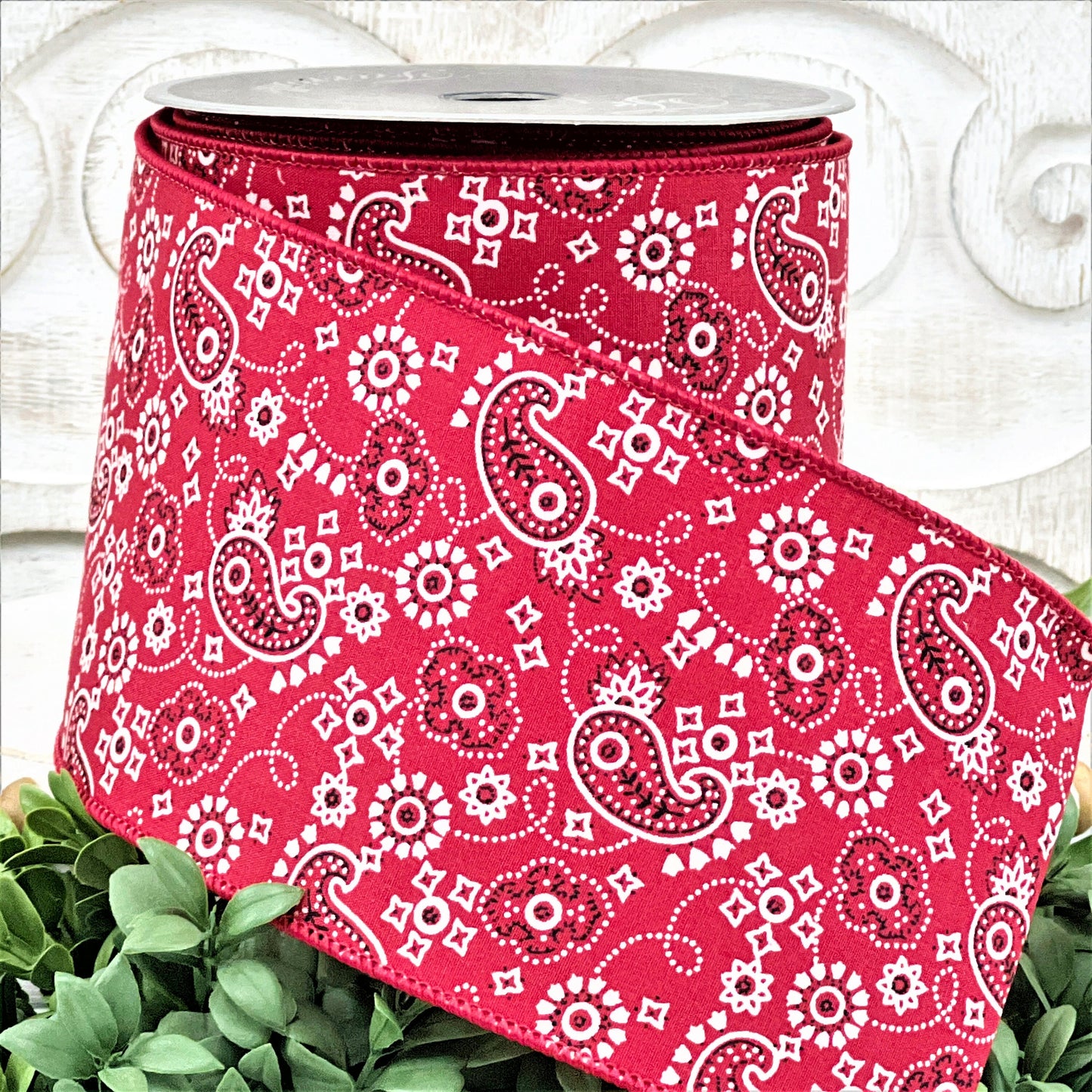 A 4&quot; roll of red paisley print ribbon sits on a white wood pedestal surrounded by greenery.