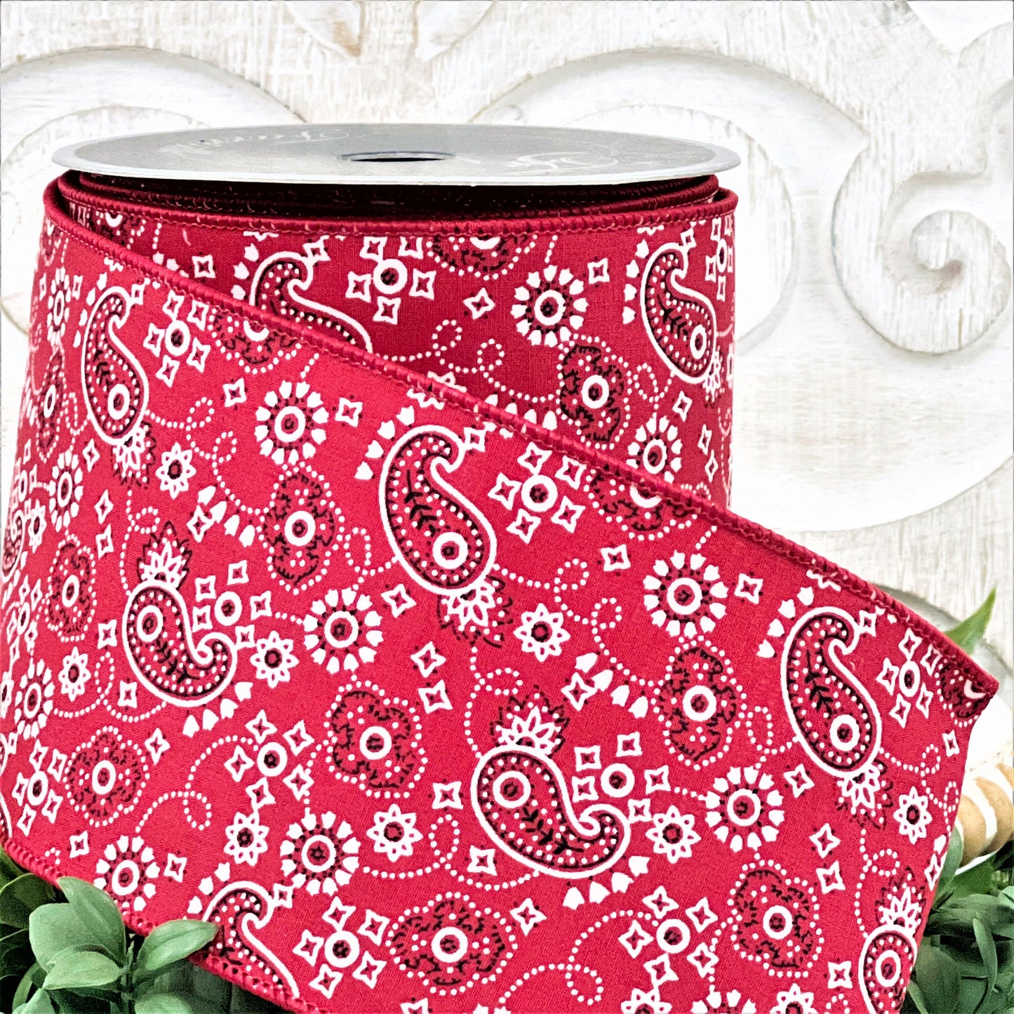 A 4&quot; roll of red paisley print ribbon sits on a white wood pedestal surrounded by greenery.