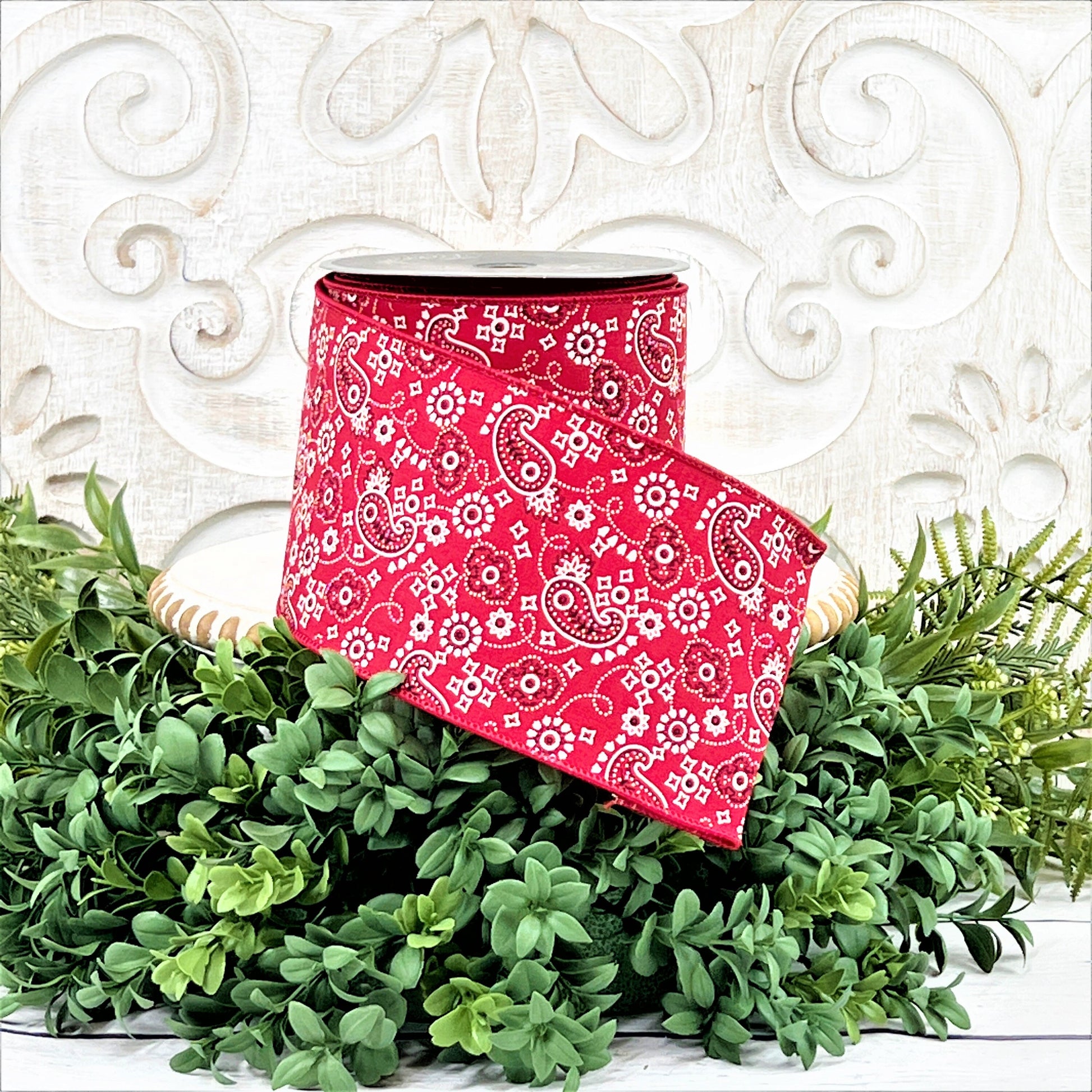 A 4&quot; roll of red paisley print ribbon sits on a white wood pedestal surrounded by greenery.