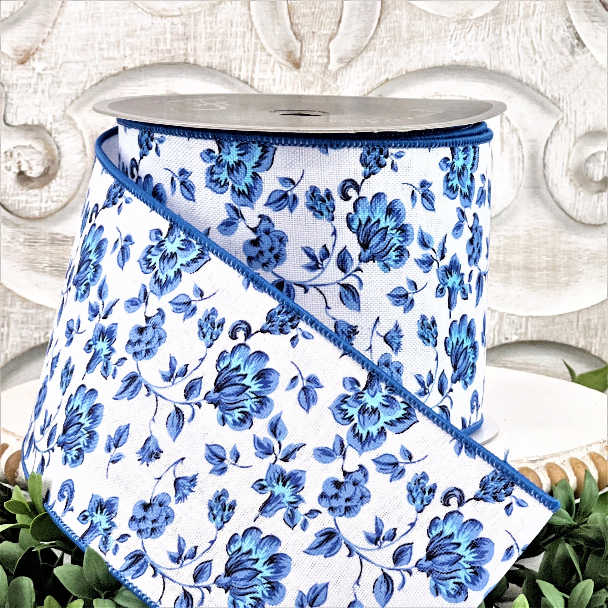 A 4&quot; roll of blue and white Dutch Porcelan flowered ribbon sits on top of a white wood pedestal surrounded by greenery.