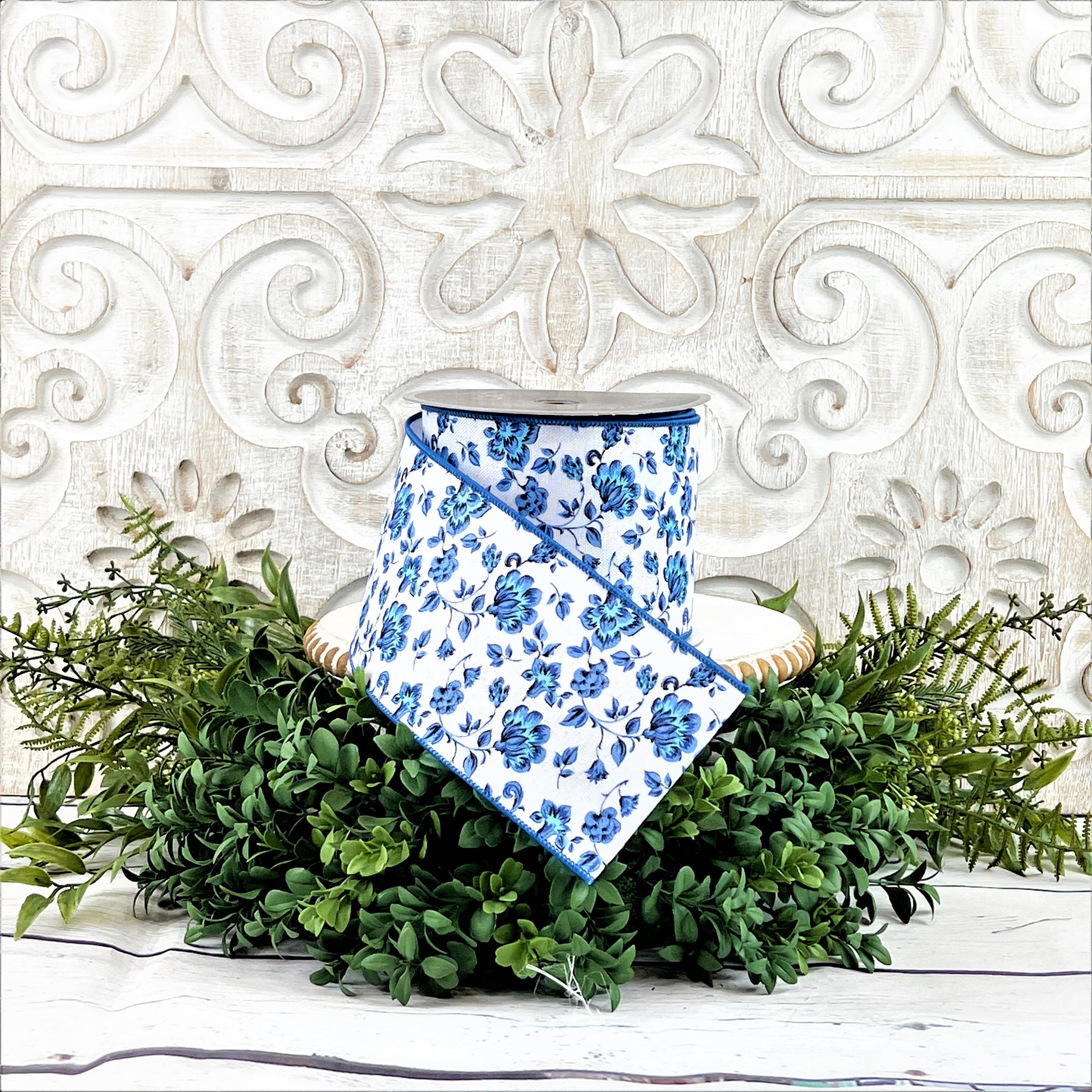 A 4&quot; roll of blue and white Dutch Porcelan flowered ribbon sits on top of a white wood pedestal surrounded by greenery.
