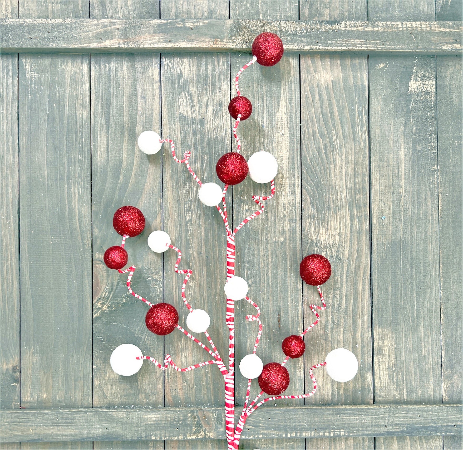 A 31 inch peppermint glitter ball spray rests against a distressed wood fence.